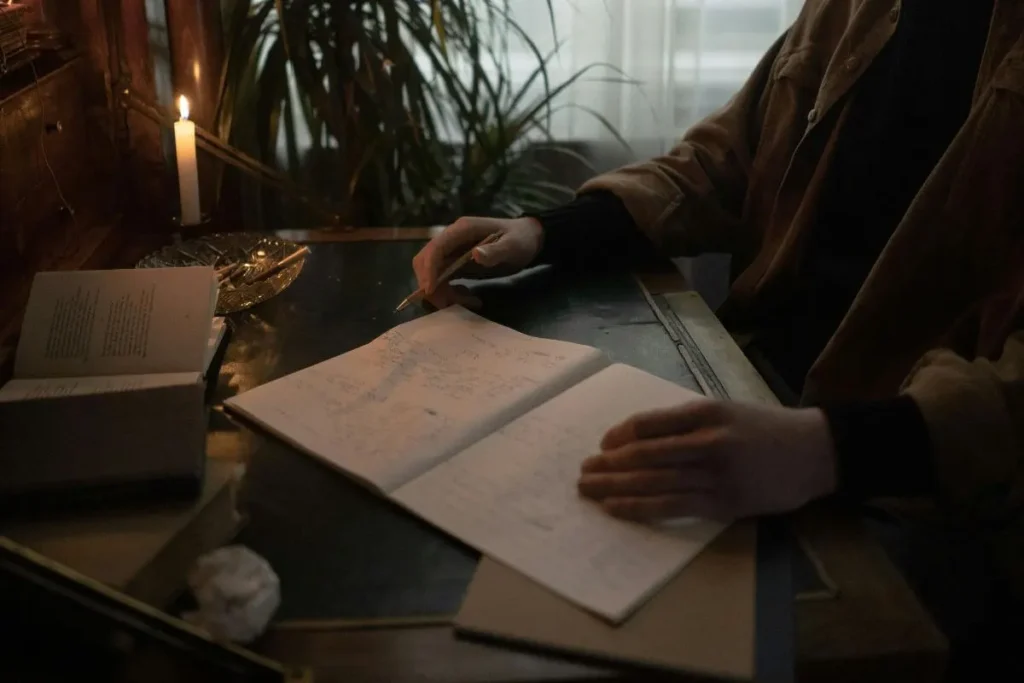 Mãos escrevendo em um caderno sobre a mesa, ao lado de uma vela acesa, em ambiente acolhedor e intimista.