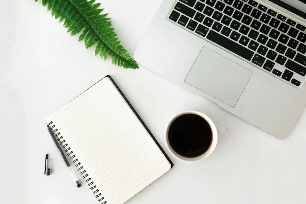 Mesa de trabalho minimalista com caderno aberto, caneta, xícara de café, notebook e folha verde sobre fundo branco.