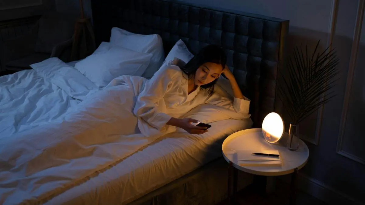 Mulher deitada na cama de olhos abertos no escuro, usando o celular ao lado de um abajur com luz suave, transmitindo inquietação e mente acelerada.