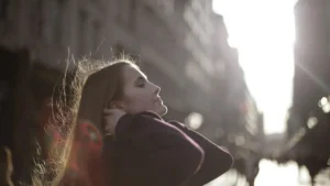 Mulher com os olhos fechados e as mãos próximas ao pescoço, em ambiente externo com luz natural suave, transmitindo calma e introspecção.