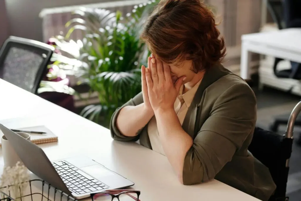 Mulher sentada à mesa com notebook, cobrindo o rosto com as mãos, demonstrando estresse, cansaço mental e sobrecarga emocional no trabalho.