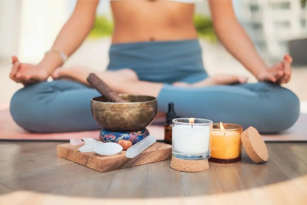Mulher sentada em postura de meditação, com as mãos abertas sobre os joelhos, próxima a velas acesas e objetos de relaxamento.