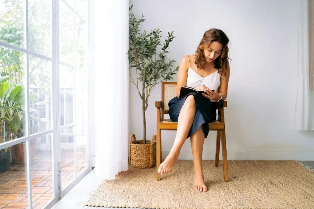 Mulher sentada em cadeira escrevendo em caderno junto à janela com plantas ao fundo e expressão concentrada e serena em momento de reconexão interna