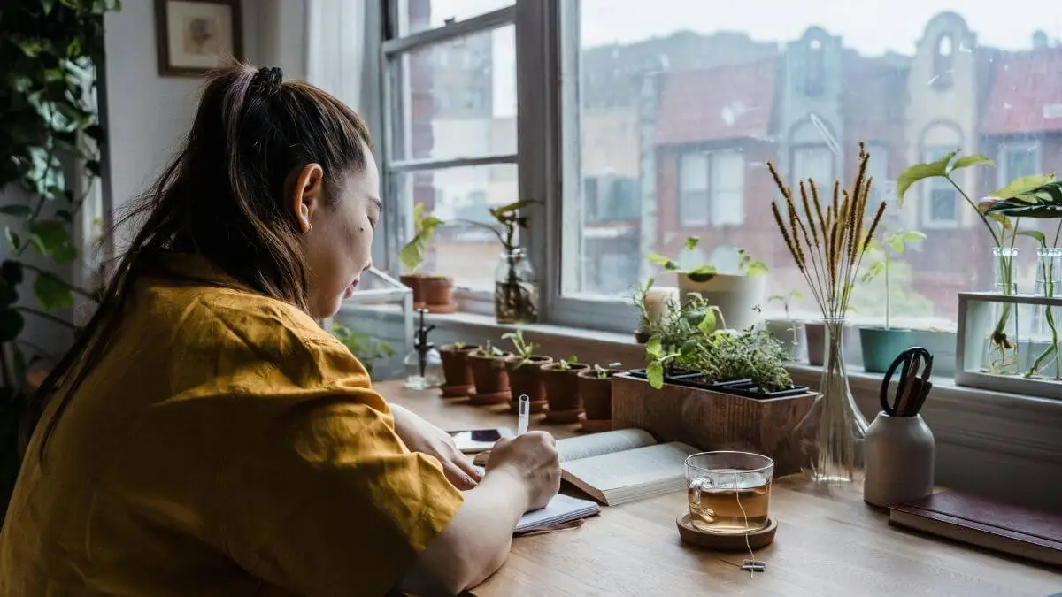 Mulher escrevendo em caderno junto à janela com plantas ao redor e chá na mesa em momento de reflexão consciente