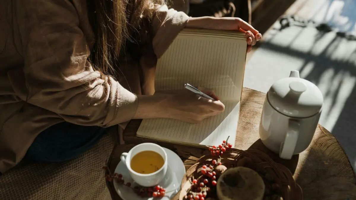 Mulher escrevendo em caderno aberto ao lado de uma xícara de chá, em ambiente aconchegante com luz natural suave.