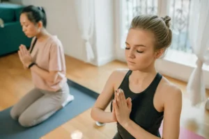 Mulher com olhos fechados em postura serena de meditação, com as mãos unidas próximas ao peito, em ambiente iluminado por luz suave.
