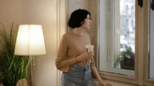 Mulher segurando um copo e olhando pela janela em ambiente acolhedor, com luz suave e expressão serena.