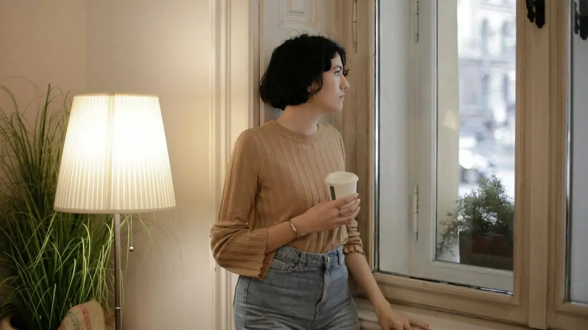 Mulher segurando um copo e olhando pela janela em ambiente acolhedor, com luz suave e expressão serena.