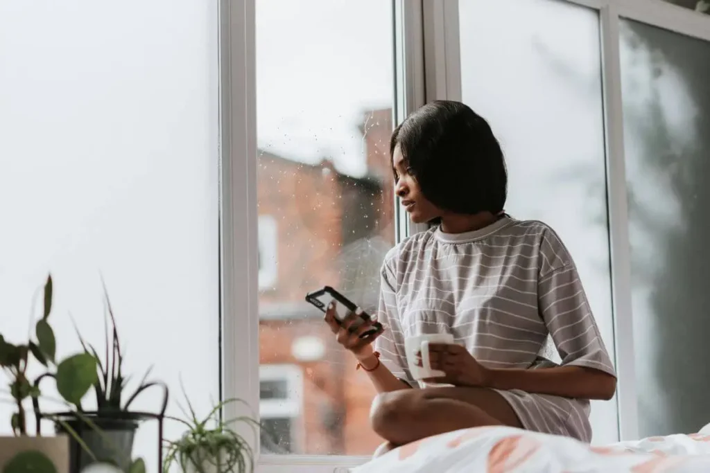 Mulher sentada perto da janela segurando uma xícara e um celular, em momento de pausa consciente com luz natural suave.