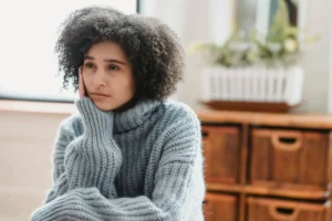 Mulher sentada sozinha em casa, com expressão reflexiva e olhar distante, transmitindo introspecção e peso emocional.