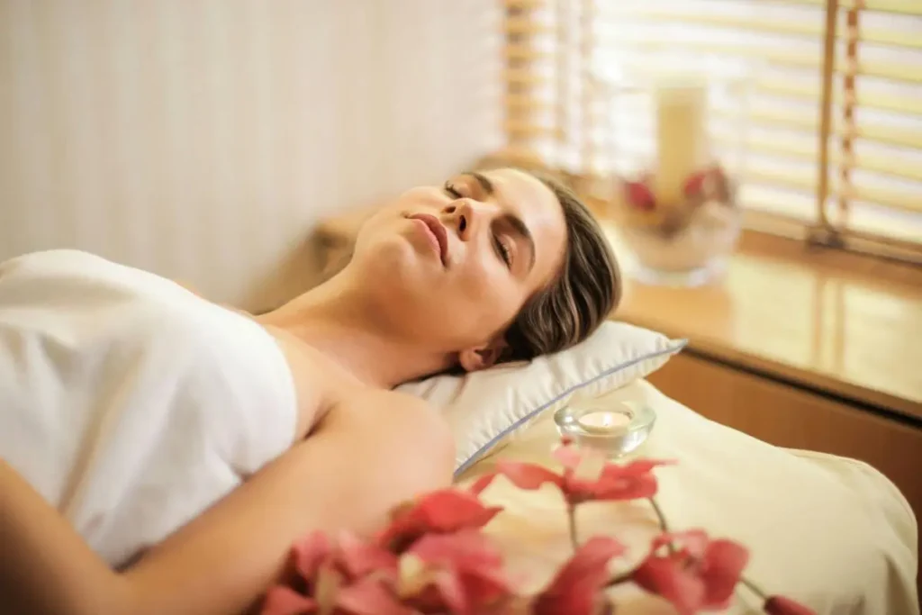 Mulher deitada em postura relaxada, com olhos fechados e expressão serena, ao lado de vela acesa em ambiente acolhedor com luz quente.