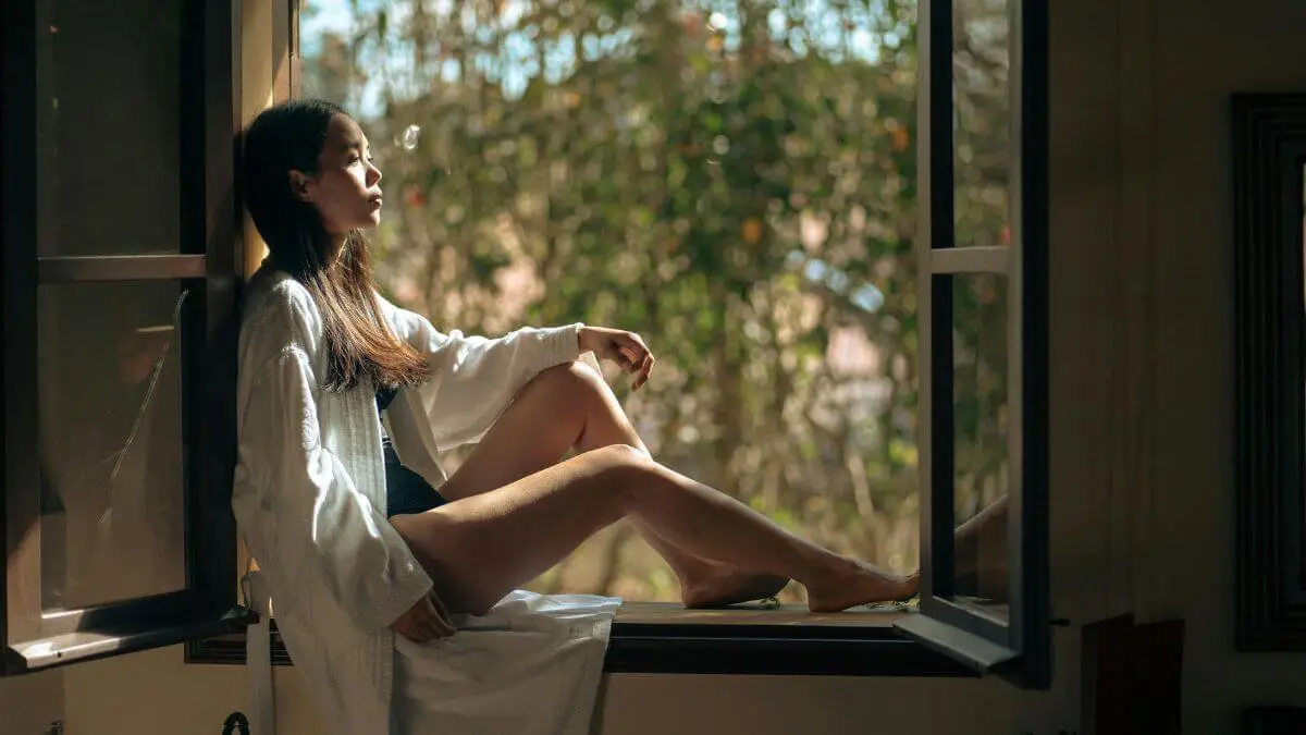 Mulher sentada no parapeito da janela com olhar introspectivo e sereno em momento de solidão emocional com luz natural suave