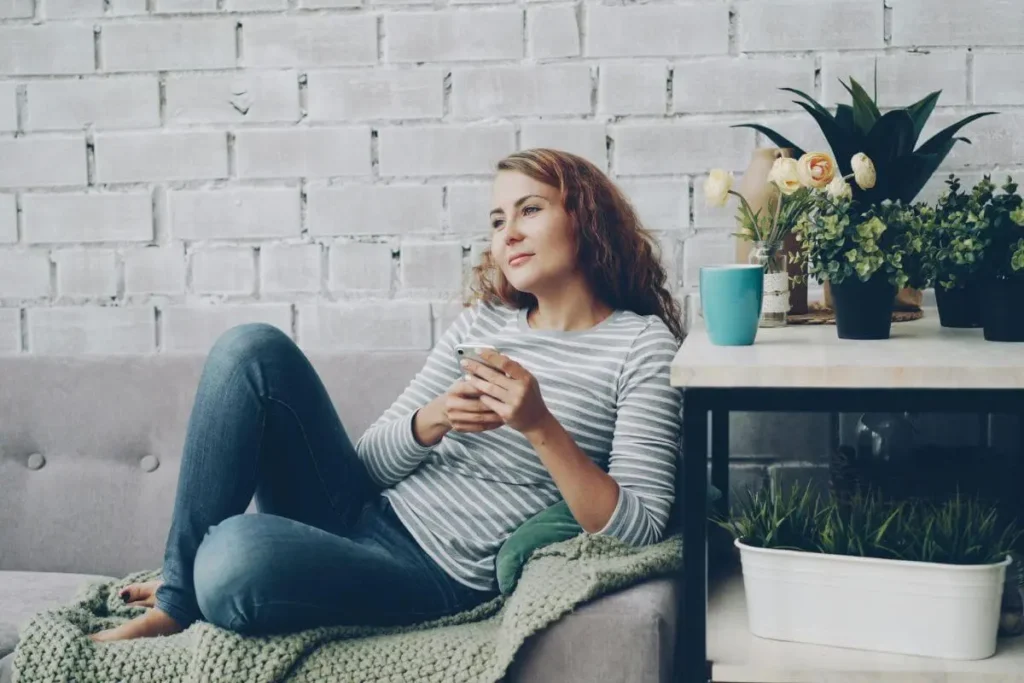 Mulher sentada no sofá em ambiente doméstico acolhedor, segurando o celular e olhando ao longe com expressão distante e pensativa.