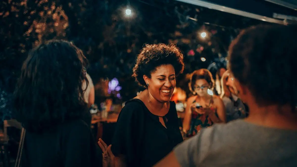 Mulher sorrindo durante uma conversa em ambiente social, com outras pessoas ao redor e iluminação quente ao fundo.