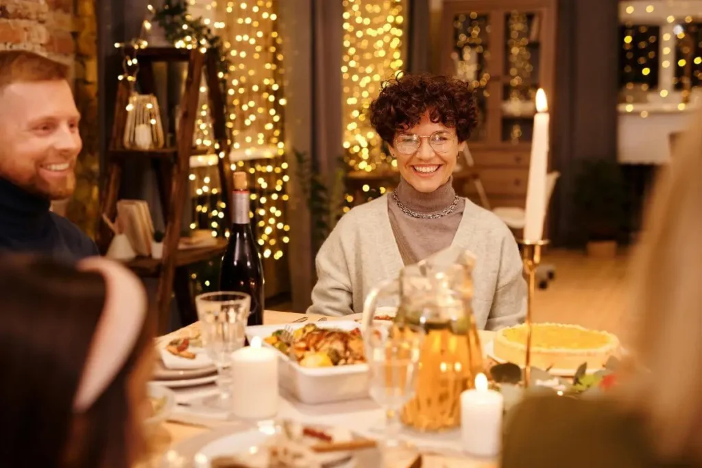 Mulher sorrindo durante um jantar em ambiente social aconchegante, com luzes quentes ao fundo e pessoas ao redor.