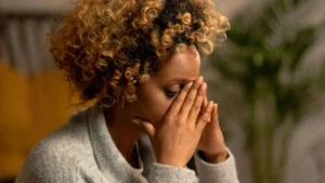 Mulher com expressão de tensão emocional, cobrindo o rosto com as mãos em ambiente doméstico com luz suave.