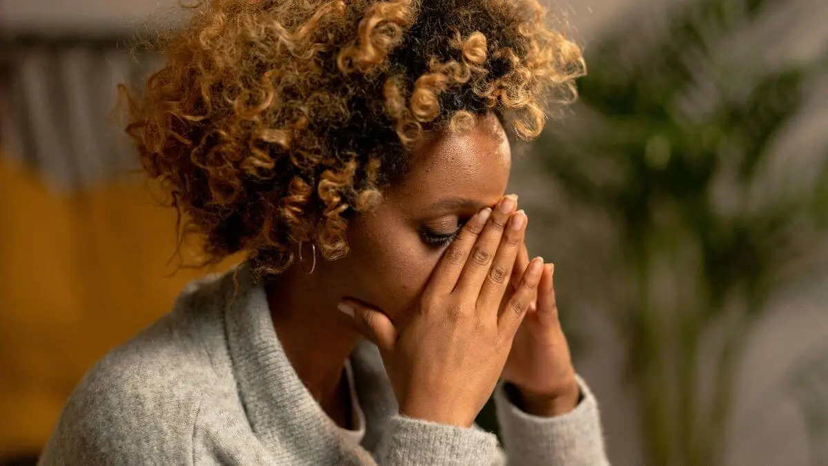 Mulher com expressão de tensão emocional, cobrindo o rosto com as mãos em ambiente doméstico com luz suave.
