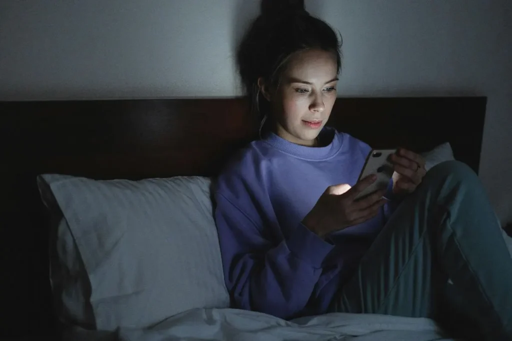 Mulher usando celular na cama à noite, com rosto iluminado pela tela e sensação de cansaço antes de dormir.