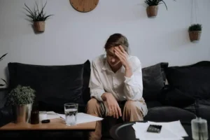 Mulher sentada no sofá com a mão na testa, em ambiente doméstico, expressando cansaço emocional e sobrecarga.