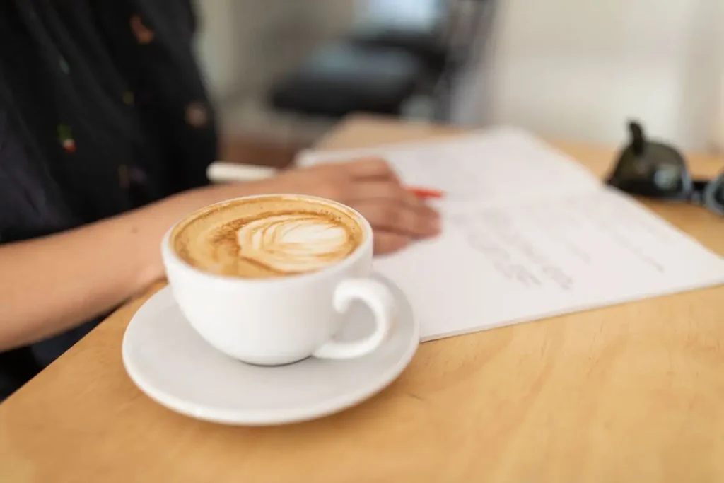 Pessoa escrevendo em um caderno ao lado de uma xícara de café, em momento de concentração, reflexão e autoconhecimento.