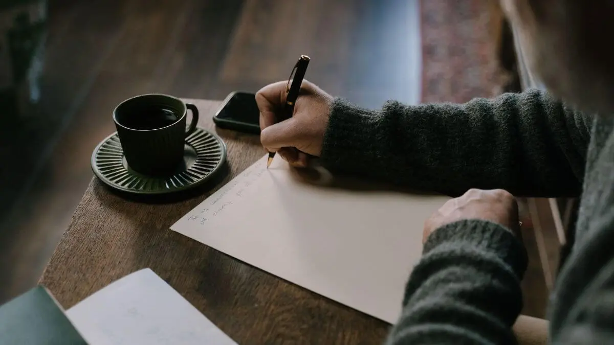 Pessoa escrevendo em uma folha ao lado de uma xícara de café, em momento de reflexão, concentração e autoconhecimento.