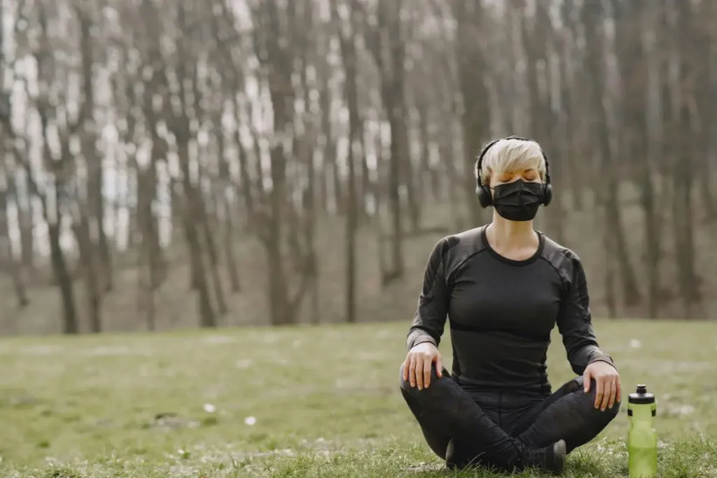 Mulher meditando sentada na grama ao ar livre, usando fones de ouvido e máscara, em ambiente de natureza tranquila.