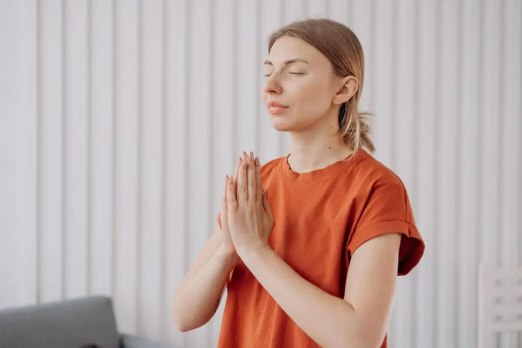 Mulher com olhos fechados e mãos unidas próximas ao peito, em momento de calma, presença e acolhimento interno em ambiente claro e tranquilo.