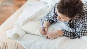 Mulher escrevendo em um caderno aberto sobre a cama, em momento de reflexão, autoconhecimento e clareza interna com luz natural suave.