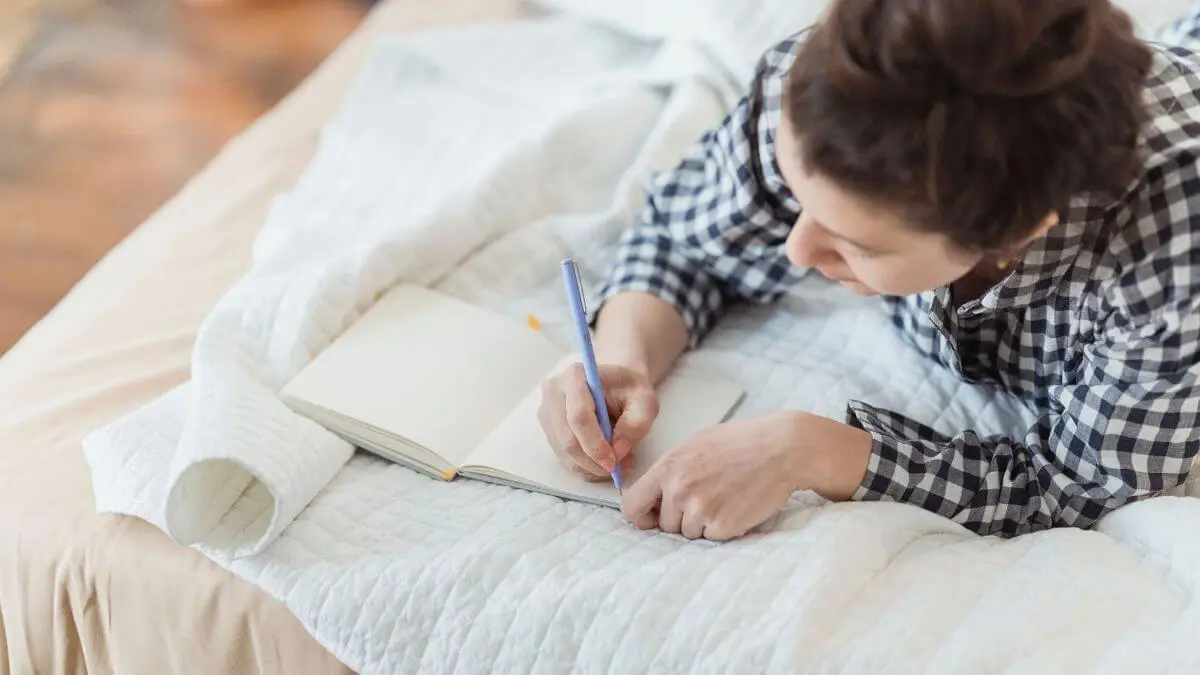 Mulher escrevendo em um caderno aberto sobre a cama, em momento de reflexão, autoconhecimento e clareza interna com luz natural suave.