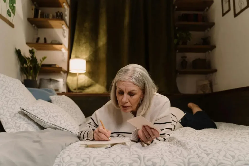 Mulher deitada em ambiente aconchegante escrevendo em um caderno, com luz quente ao fundo, em momento de reflexão, autocuidado e prática consciente.