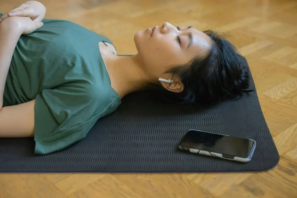 Mulher deitada em tapete de exercícios usando fones de ouvido, praticando meditação guiada com expressão serena.
