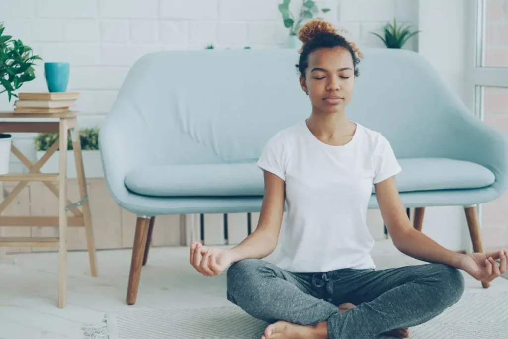 Mulher sentada em posição de meditação com os olhos fechados, praticando respiração consciente em ambiente claro, tranquilo e acolhedor.