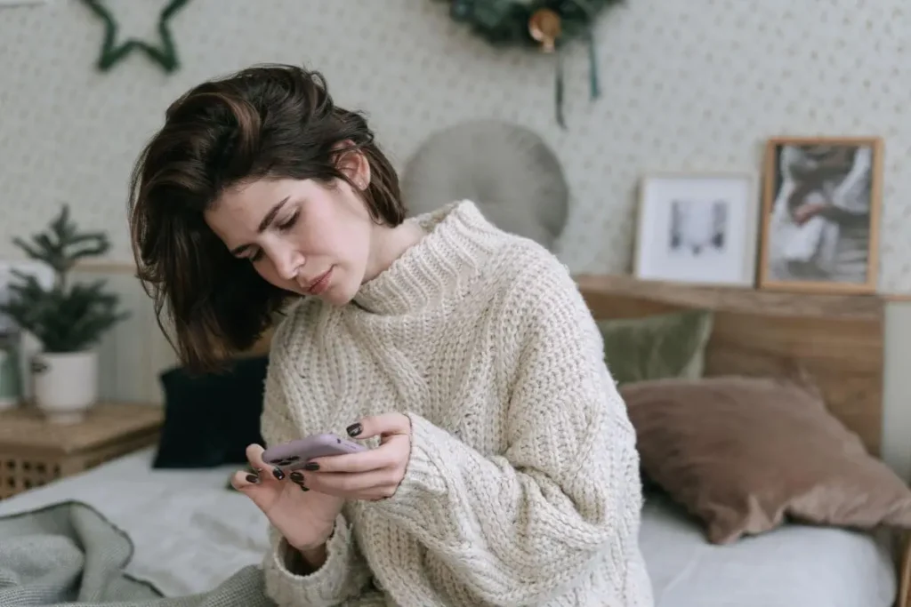 Mulher sentada no quarto olhando para o celular com expressão séria e desconfortável, transmitindo comparação, tristeza e incômodo emocional.