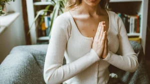 Mulher sentada no sofá com as mãos unidas em posição de calma, praticando respiração consciente em ambiente acolhedor com luz natural.
