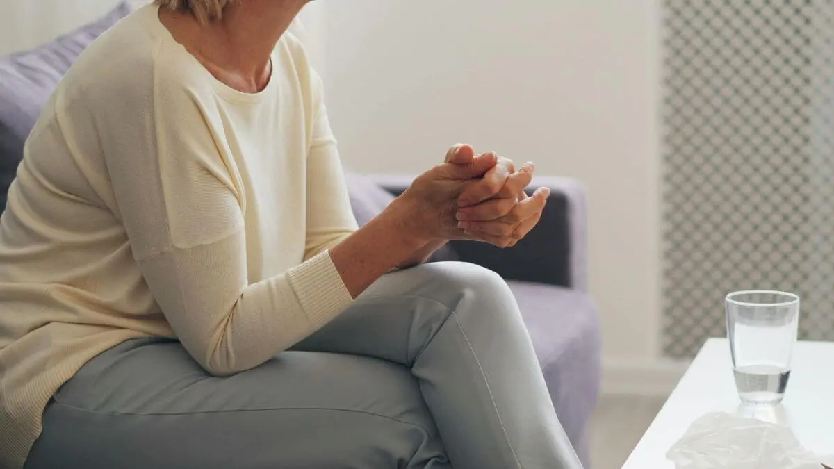 Mulher sentada em ambiente claro, com as mãos unidas, copo de água e lenço sobre a mesa, transmitindo cuidado, acolhimento e bem-estar.
