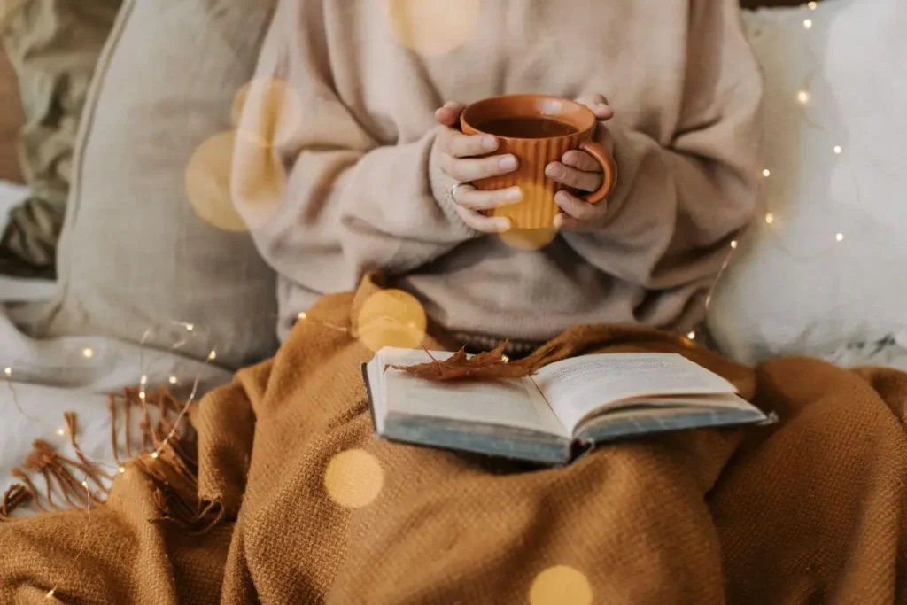 Mulher segurando uma xícara de chá enquanto lê um livro em ambiente aconchegante com luzes suaves.